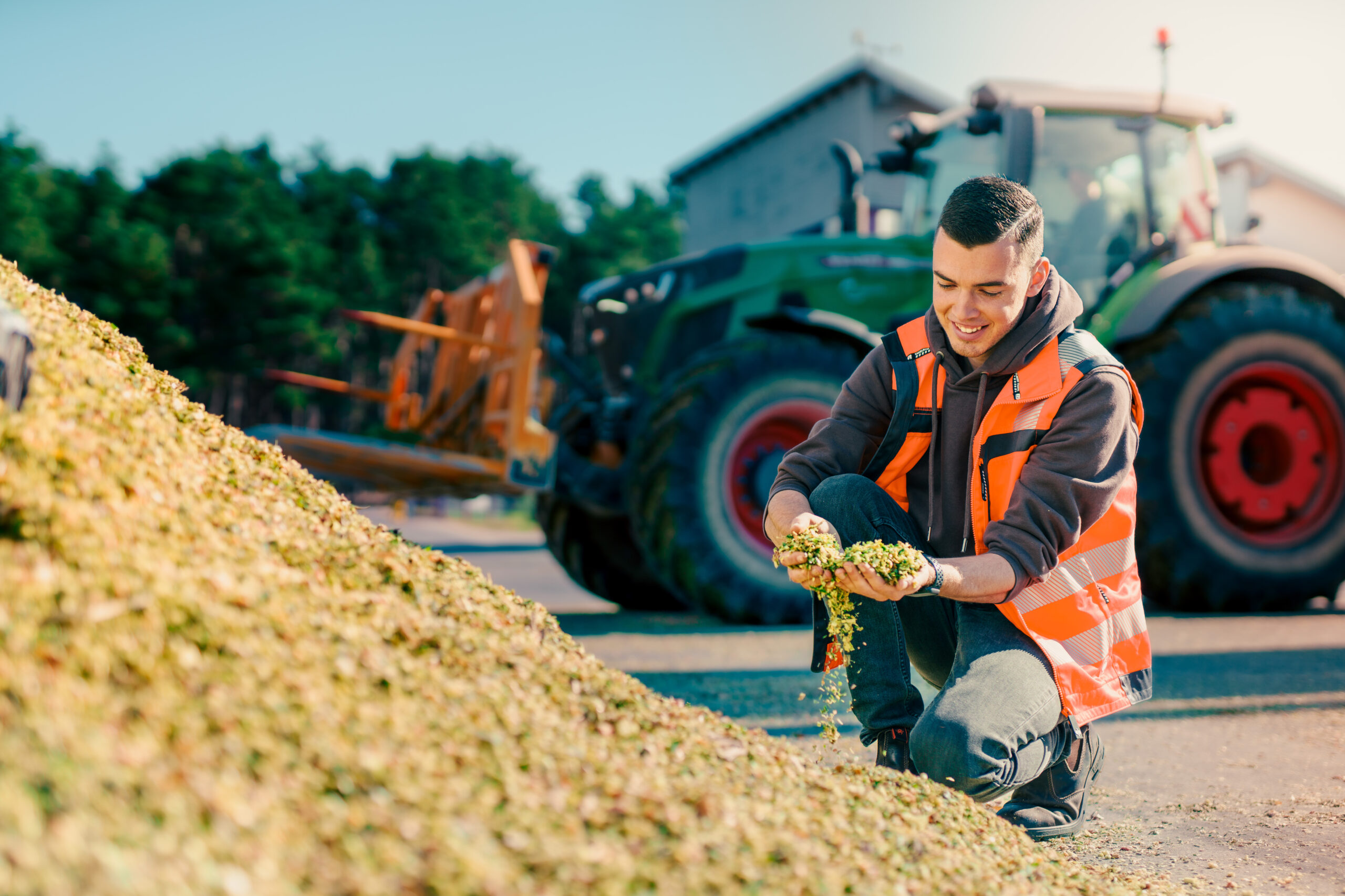 Junger Mann mit Warnweste hockt am Boden und entnimmt von einem Haufen Substrat eine Handvoll. Im HIntergrund steht ein Radlader.